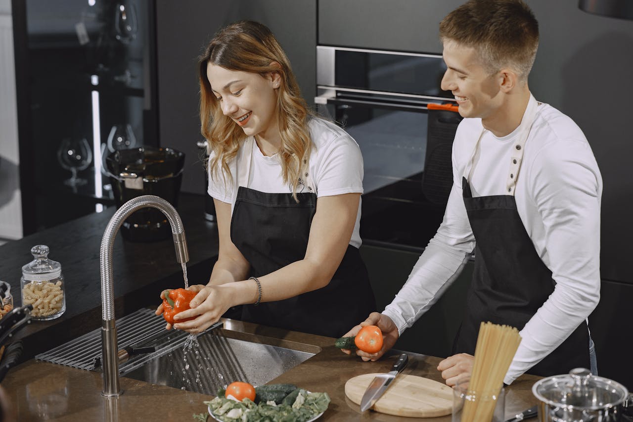 A smiling couple washes vegetables in a sleek kitchen, preparing a meal together.