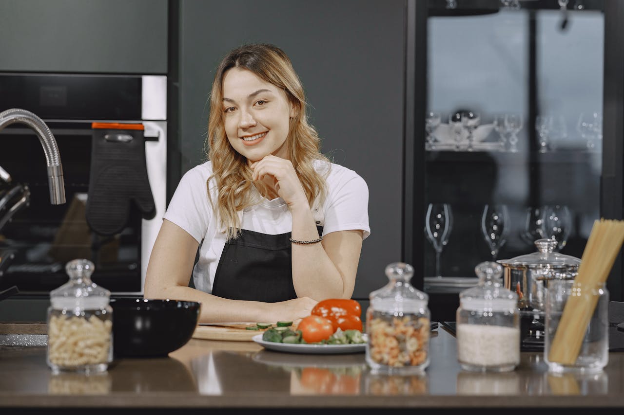A smiling woman in an apron preparing food in a modern kitchen setting.